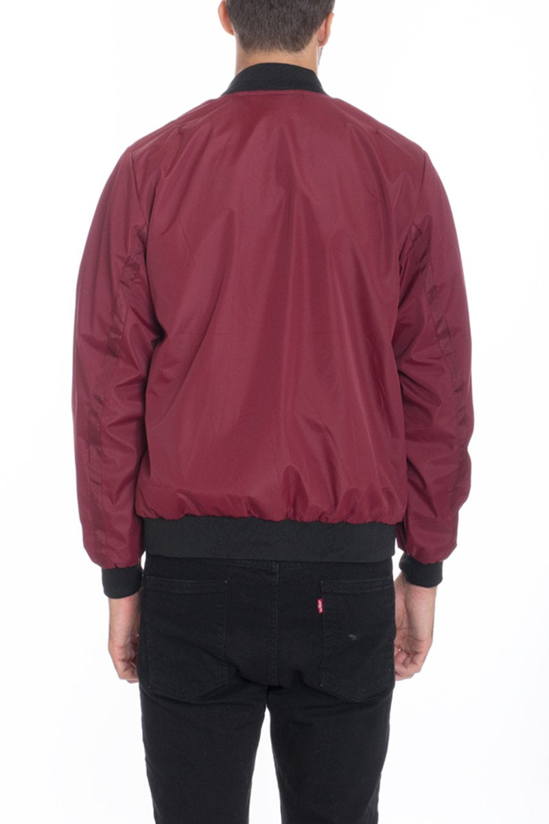 A man stands with his back to the camera against a white background, wearing the Lime Milo Mens Vinyl Waterproof Windbreaker in maroon with black cuffs and collar, paired with black jeans.