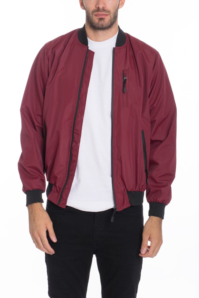 A man stands against a plain white background wearing the Lime Milo Mens Vinyl Waterproof Windbreaker in maroon with black cuffs and collar, styled over a white t-shirt and black pants. The windbreaker features a zippered chest pocket.