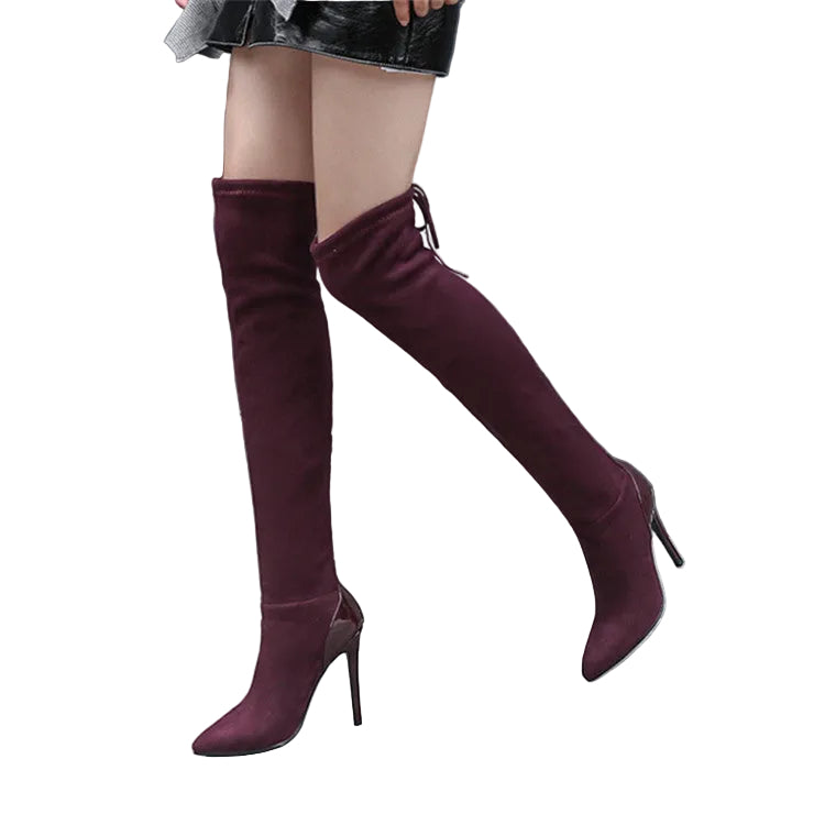 A person wearing over-the-knee pointed-toe stiletto boots in a deep burgundy or plum tone, standing against a dark gray wall and concrete floor.