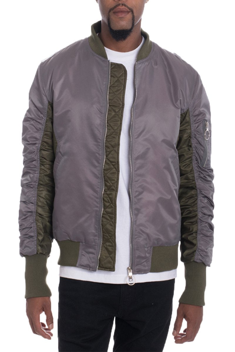 A man wearing the Lime Milo TWO TONE BOMBER in gray with olive green accents, featuring ribbed cuffs and hem, a zippered left sleeve pocket, and scrunched sleeves. He pairs it with a white T-shirt and black pants; his head is partially out of frame.