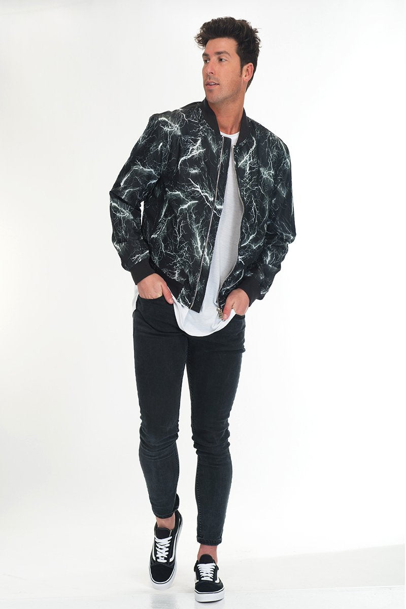 A man poses in Lime Milo's BOLT BOMBER JACKET—a black and grey jacket with a bold white lightning pattern and zipper closure—styled with a white T-shirt, black skinny jeans, and black sneakers with white soles.