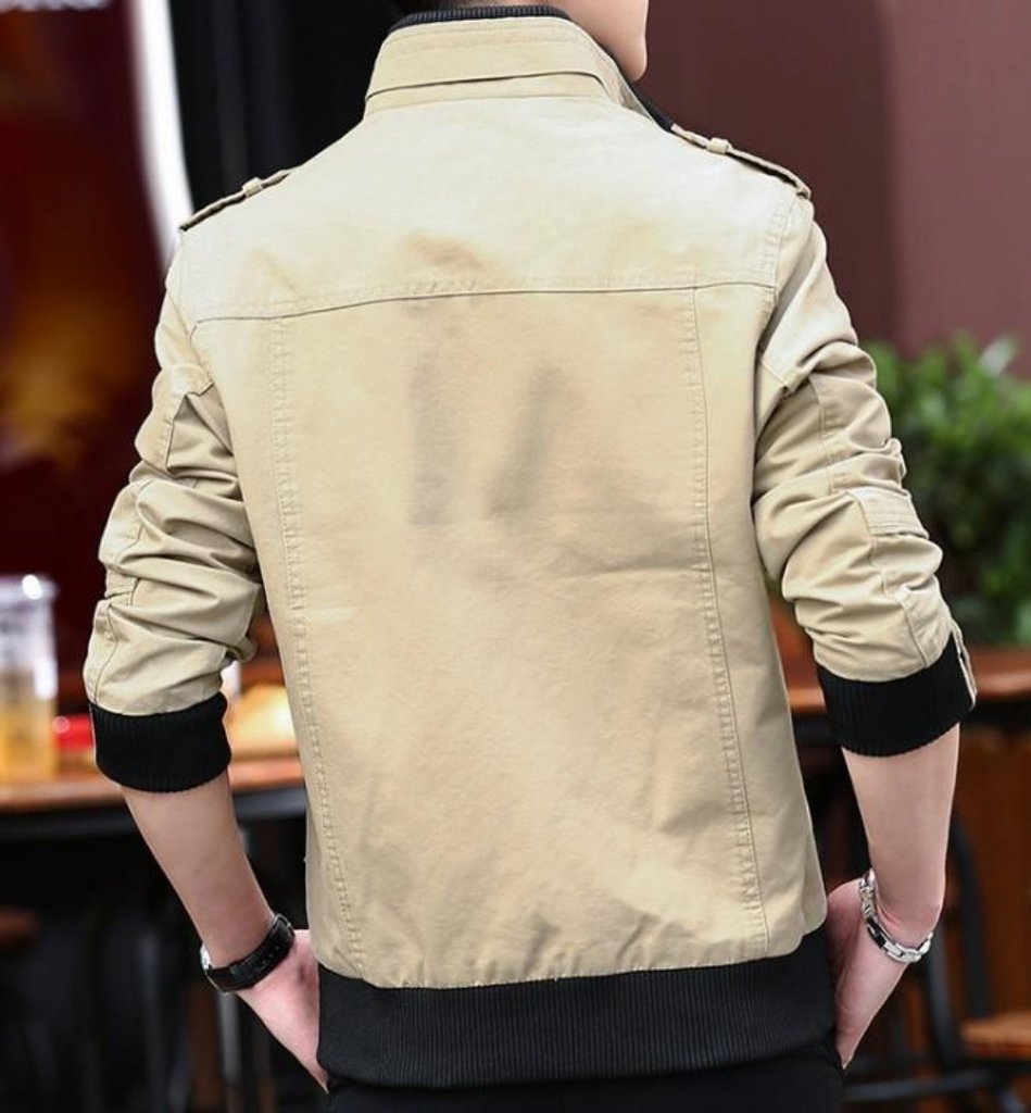 A person is shown from behind indoors, wearing the Yellow Pandora Mens Military Theme Short Jacket in light beige with black cuffs and slightly pushed-up sleeves. Blurred furniture and plants appear in the background.