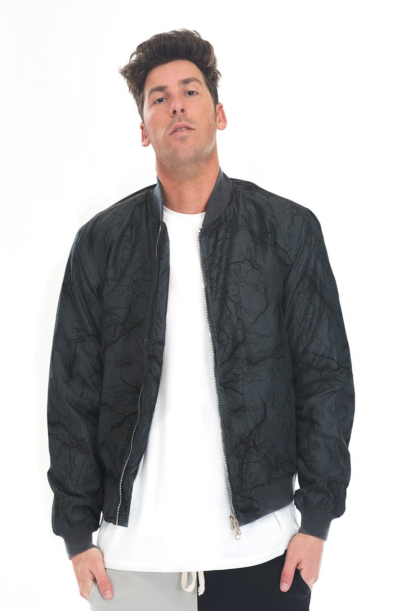 A man with short brown hair wears the Lime Milo BOLT BOMBER JACKET, featuring a zipper closure, over a long white T-shirt and black pants, standing against a plain white background with a neutral expression.