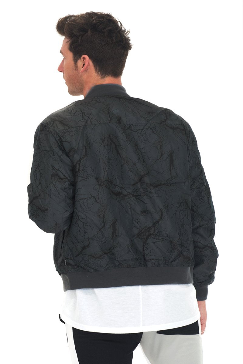 A man with dark hair, facing away, wears the Lime Milo BOLT BOMBER JACKET—a black-grey zip-up—over a white shirt and black-and-white pants against a white background.