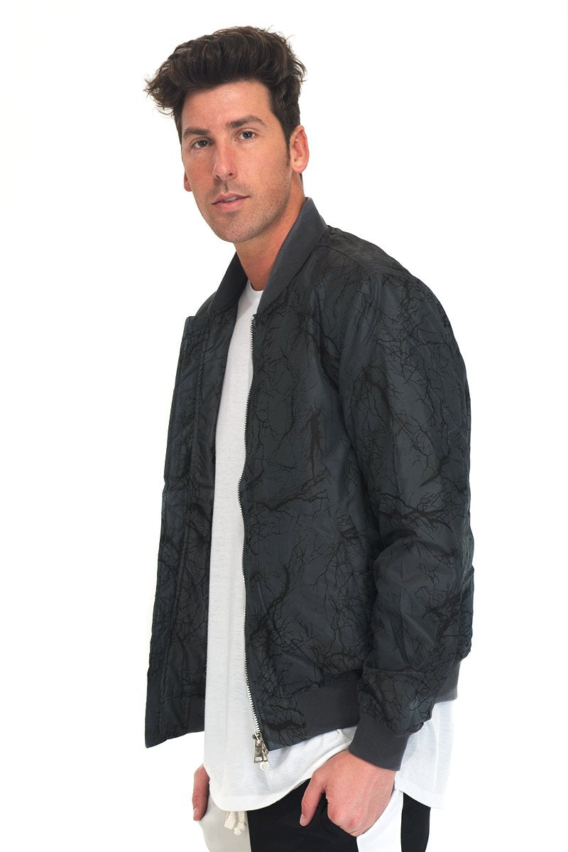 A man with short, dark hair models the Lime Milo BOLT BOMBER JACKET, a textured black-grey piece with zipper closure, over a long white t-shirt against a plain white background, looking at the camera.