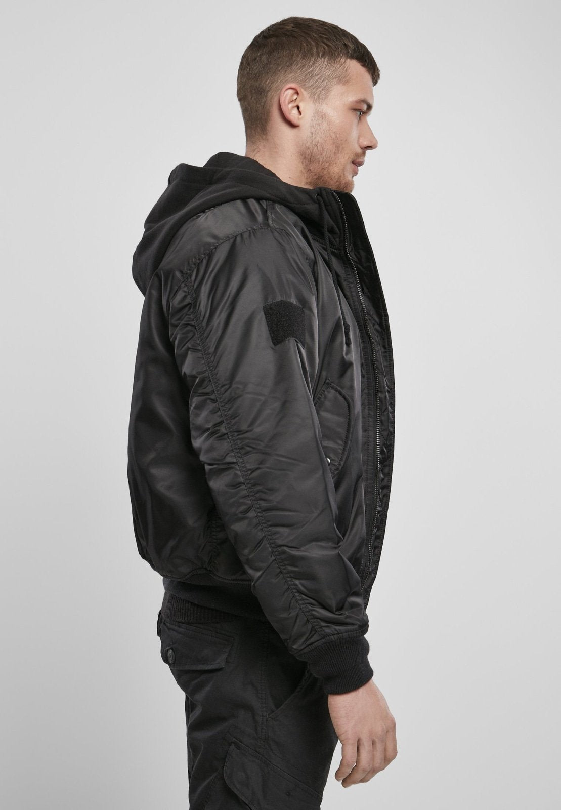 A man with short hair stands in profile against a plain light gray background, wearing the Callisto Hooded MA1 Bomber Jacket with warm lining over a black hoodie and black pants.