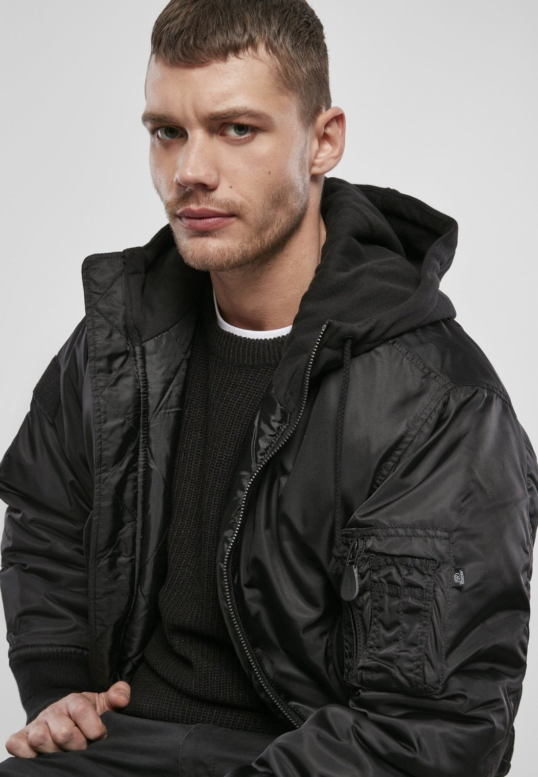 A man with short brown hair and a trimmed beard wears the Callisto Hooded MA1 Bomber Jacket over a hoodie and sweater, sitting and looking slightly to the side against a plain light background.