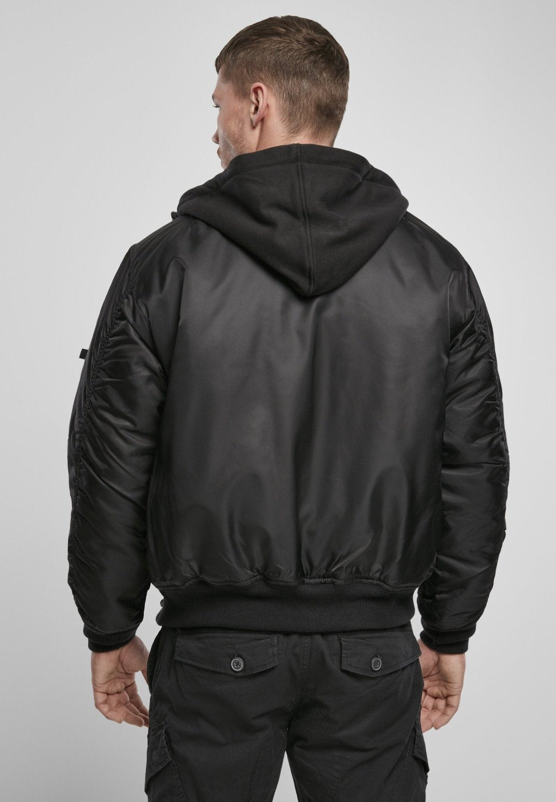 A man faces away from the camera wearing the Callisto Hooded MA1 Bomber Jacket over a black hoodie and black cargo pants, set against a plain light gray background.