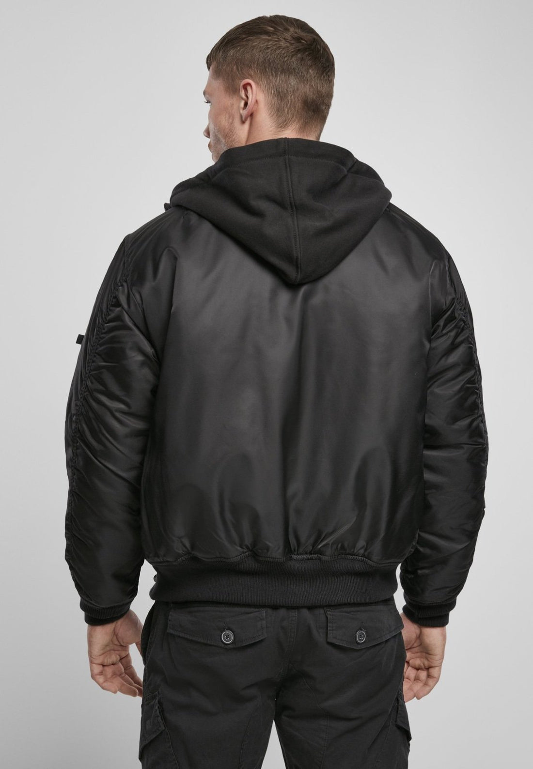 A man faces away from the camera wearing the Callisto Hooded MA1 Bomber Jacket over a black hoodie and black cargo pants, set against a plain light gray background.