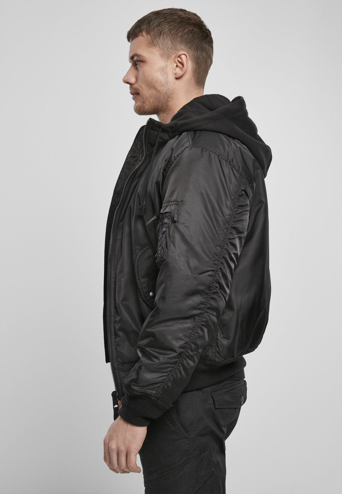 A man with short hair wears the Callisto Hooded MA1 Bomber Jacket over a black hoodie and black pants, standing in profile against a plain light gray background.