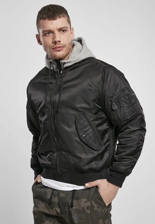 A man wears a Callisto Hooded MA1 Bomber Jacket over a gray hoodie with his hands in the pockets of wind and water repellent camouflage pants, standing against a plain light gray background.