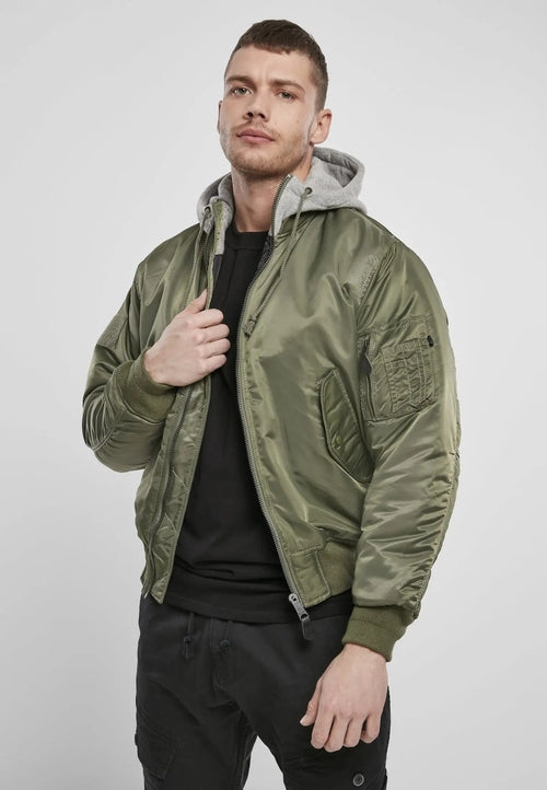 A man wears the Callisto Hooded MA1 Bomber Jacket in wind- and water-repellent olive green with warm lining, paired with a gray hoodie and black pants. He holds the collar while facing the camera against a plain light background.