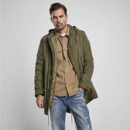 A man in a Callisto M51 US Parka Jacket, featuring an olive-green military style and removable thermal lining, is worn over a tan shirt and blue jeans as he stands against a plain light gray background, looking neutrally at the camera.